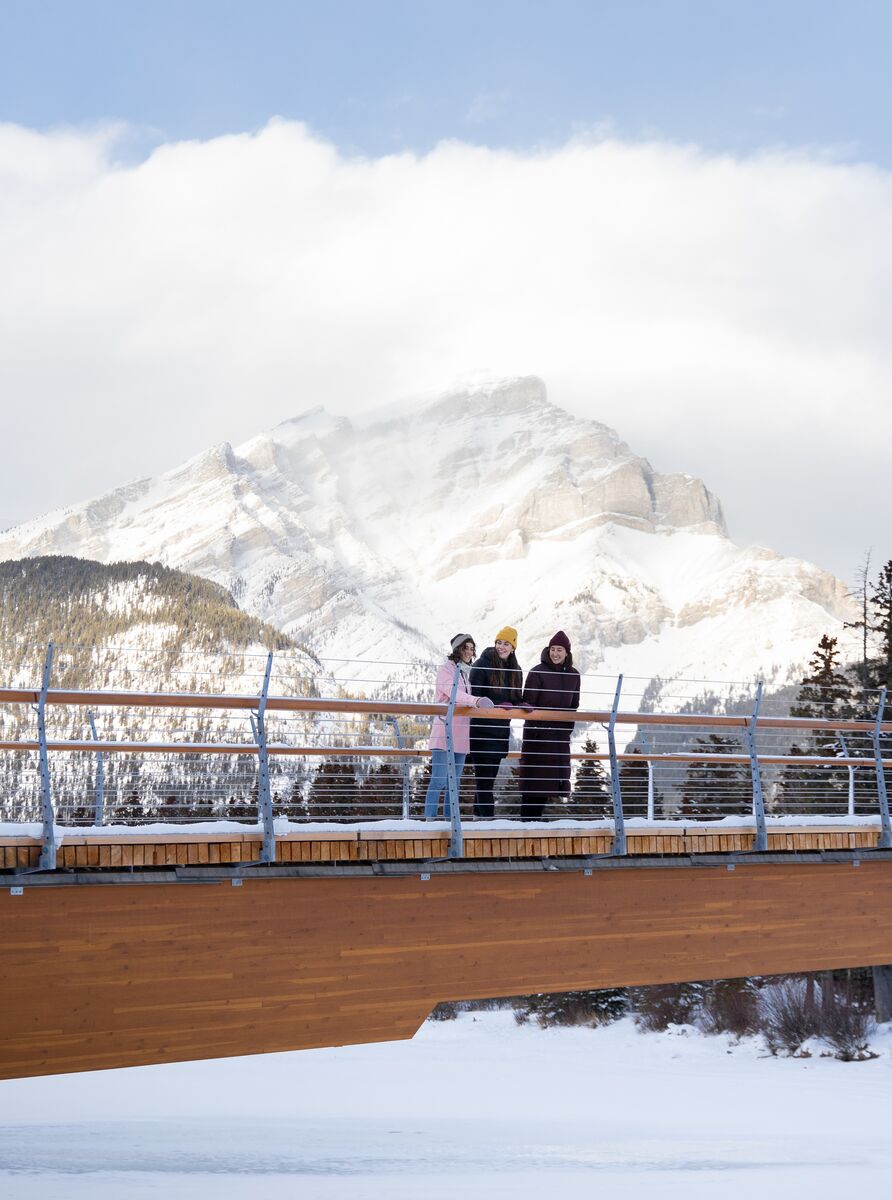 Three women stand on the Nancy Pauw bridge with Cascade Mountain behind them in Banff National Park.